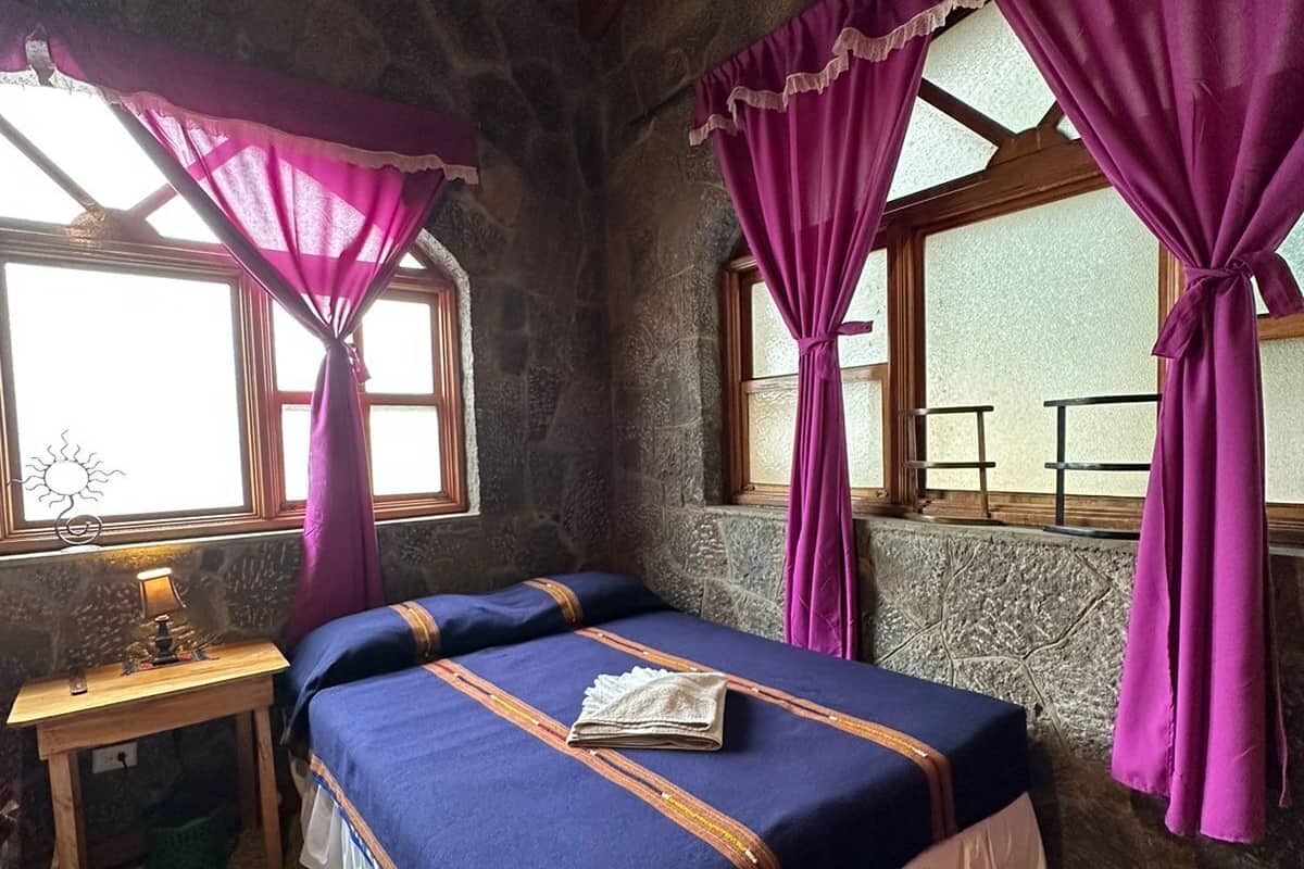 Una habitación con paredes de piedra, una cama, una mesa de madera y cortinas moradas.