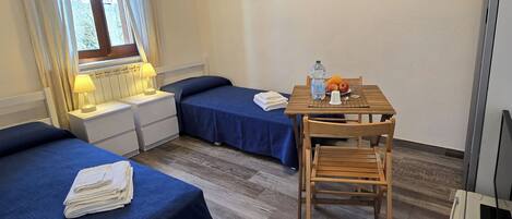 Une chambre d’hôtel avec deux lits, une petite table avec une chaise, un radiateur et une fenêtre avec des rideaux.