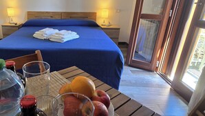 A hotel room with a large bed, two bedside lamps, a table with fruit and a bottle, and a view through a glass door. - Alghero in Bicicletta (Alghero)