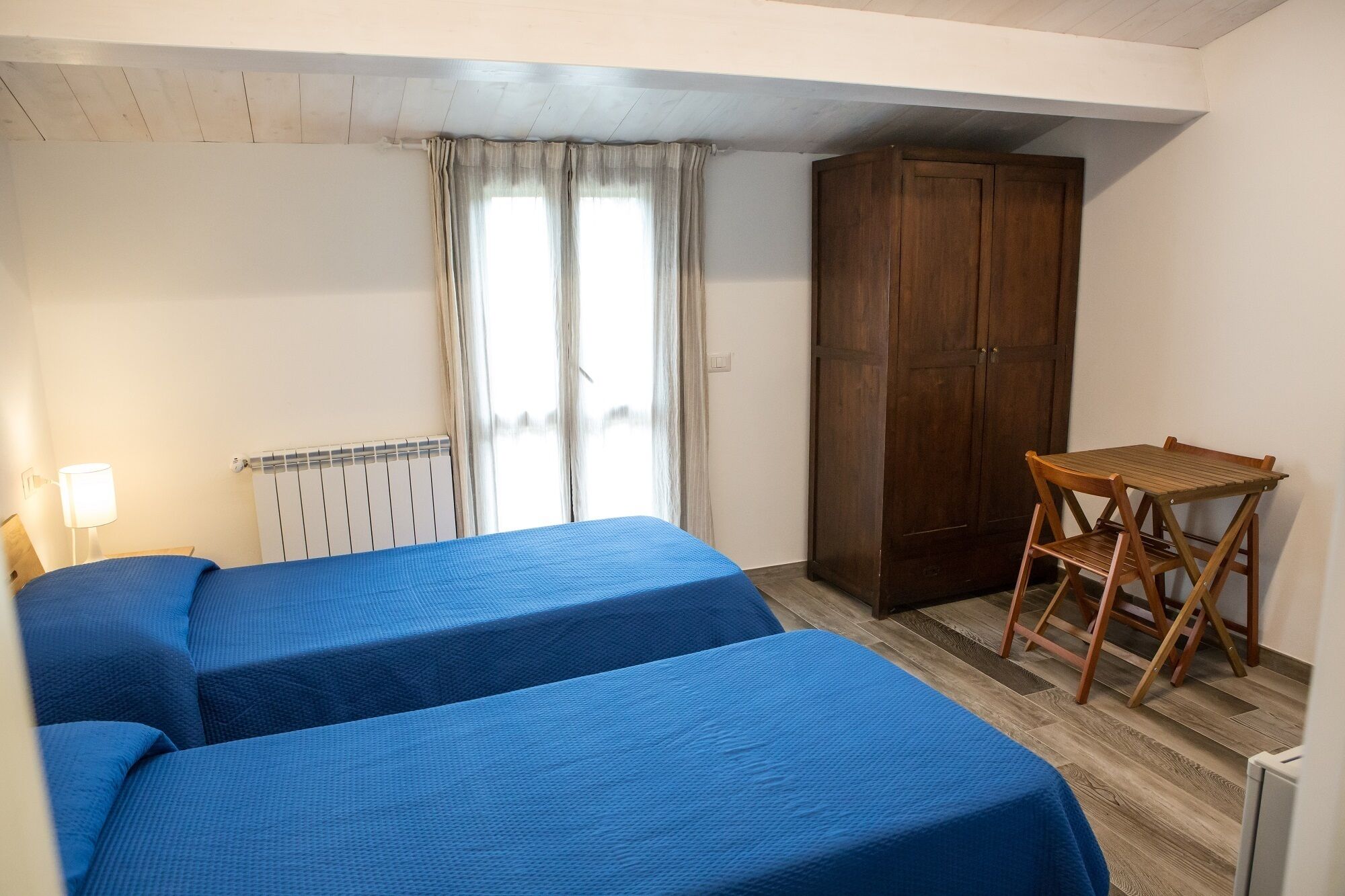 A room with two beds, a wooden desk, a chair, a radiator, and a wardrobe.