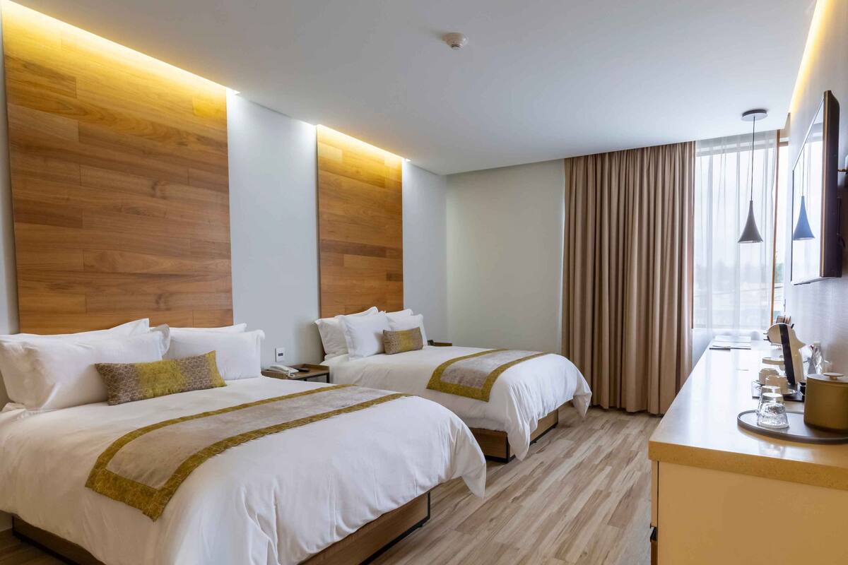 A hotel room with two beds, wooden headboards, a desk with a lamp, and a large window with curtains.