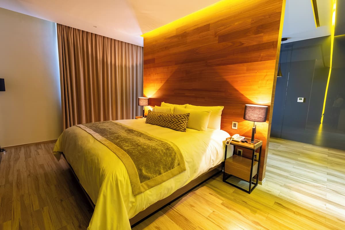 A modern hotel room with a wooden headboard, a large bed with yellow bedding, a bedside table with a lamp, and a wooden floor.