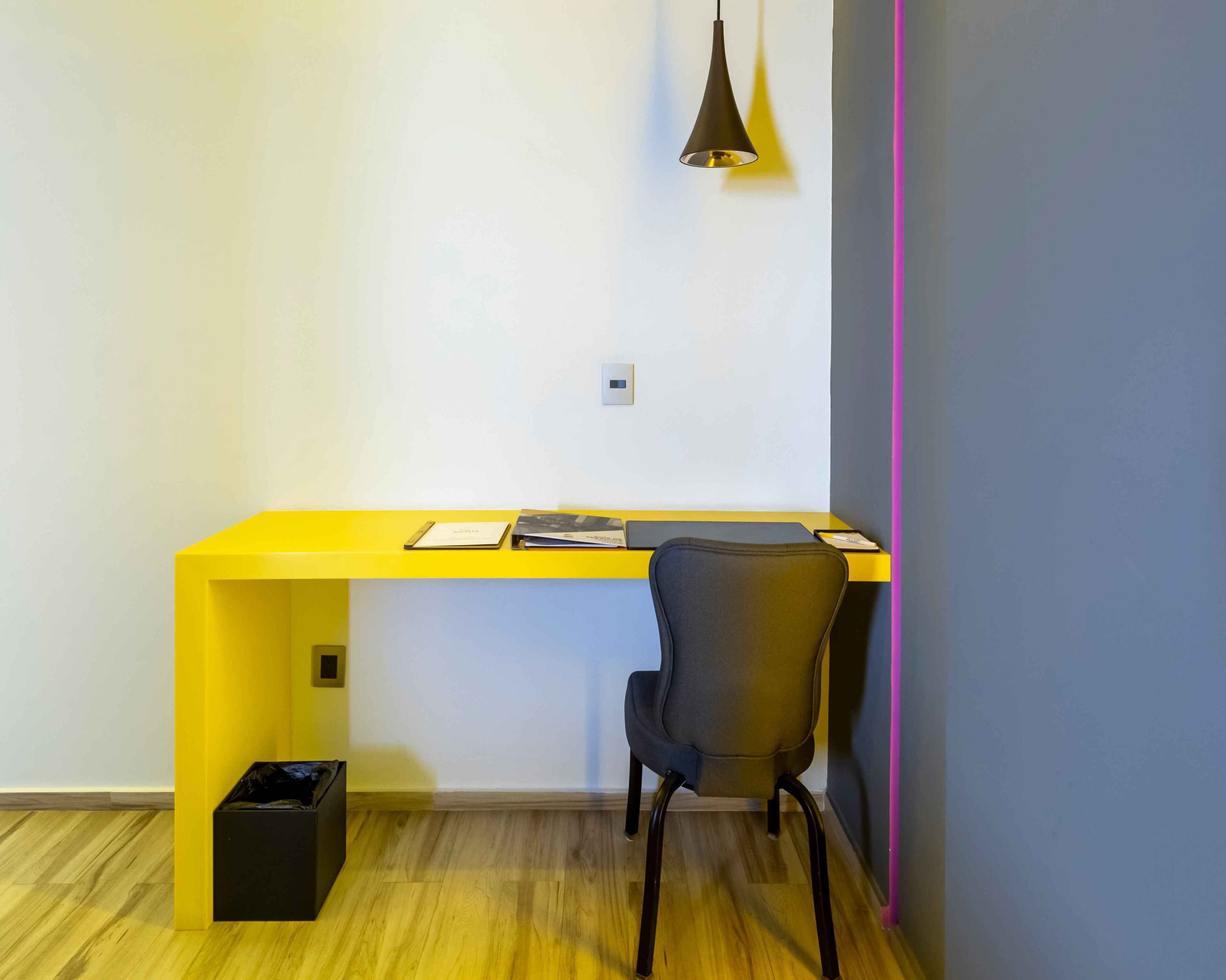 Un espacio de trabajo moderno con un escritorio amarillo, una silla negra, una lámpara colgante y un cesto de basura.