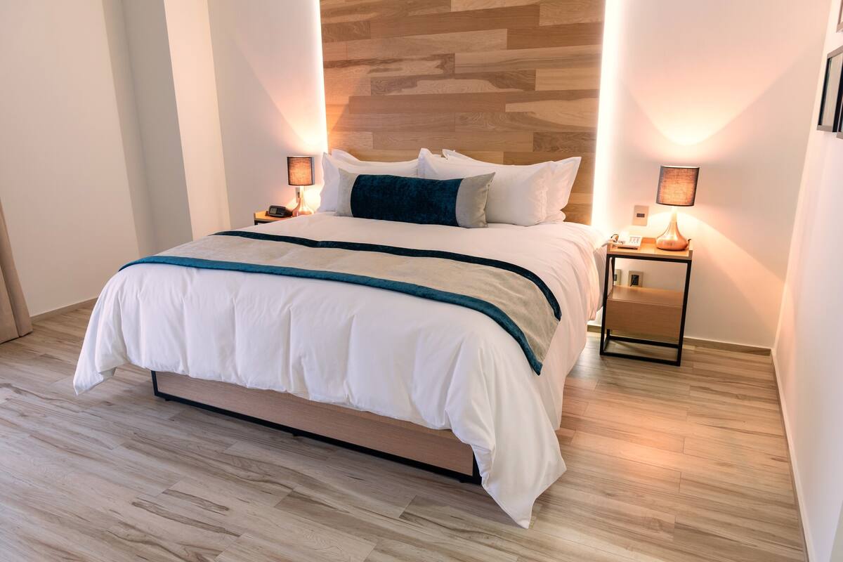 A modern hotel room with a large bed, wooden headboard, two bedside tables with lamps, and a light wooden floor.