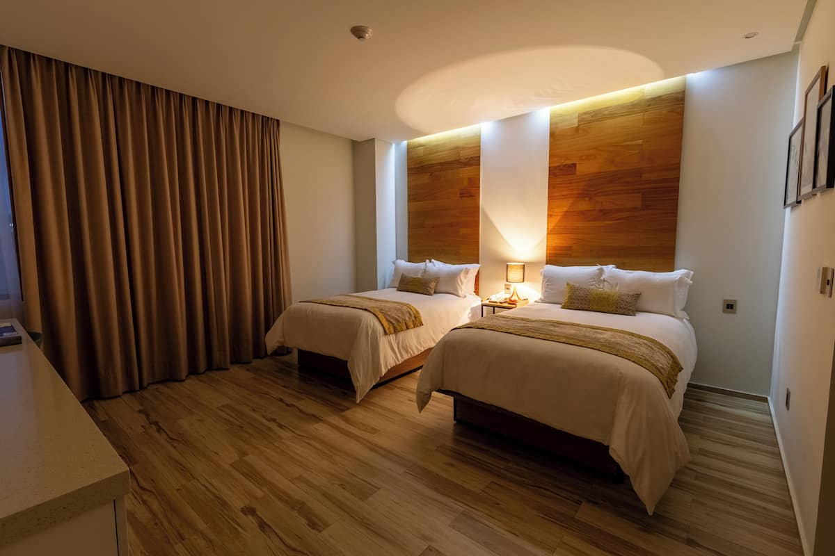 A hotel room with two beds, wooden flooring, a large window with curtains, and a wall-mounted light fixture.
