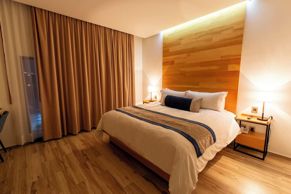 A hotel room with a large bed, wooden flooring, a nightstand with a lamp, and a view of the city through the window.