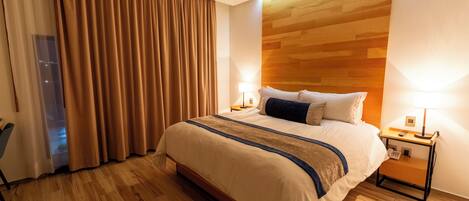 Habitación de hotel con una cama grande, suelo de madera, una mesita de noche con lámpara y vista a la ciudad desde la ventana.