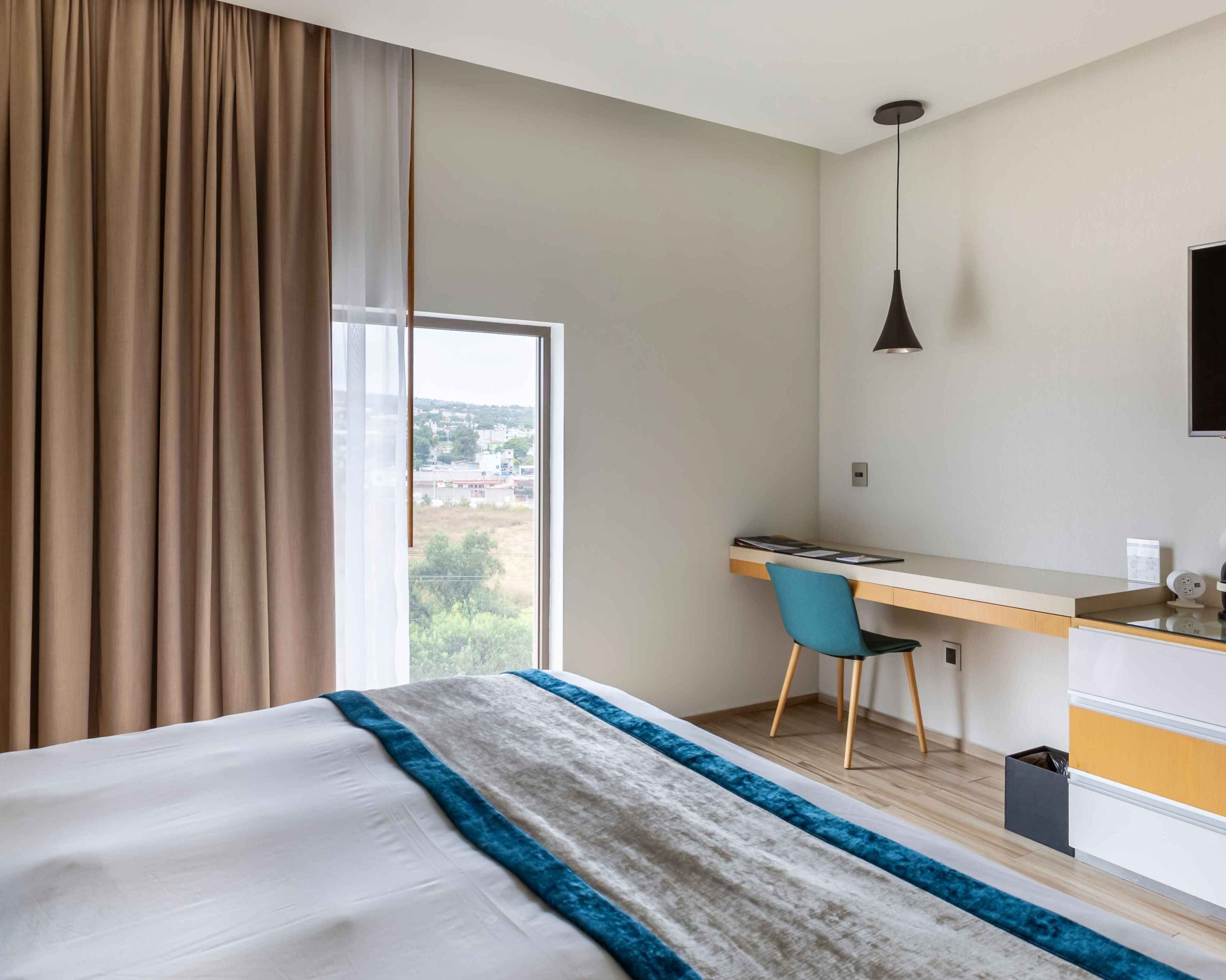Habitación de hotel con cama, escritorio, silla, ventana con cortinas y vista al exterior.