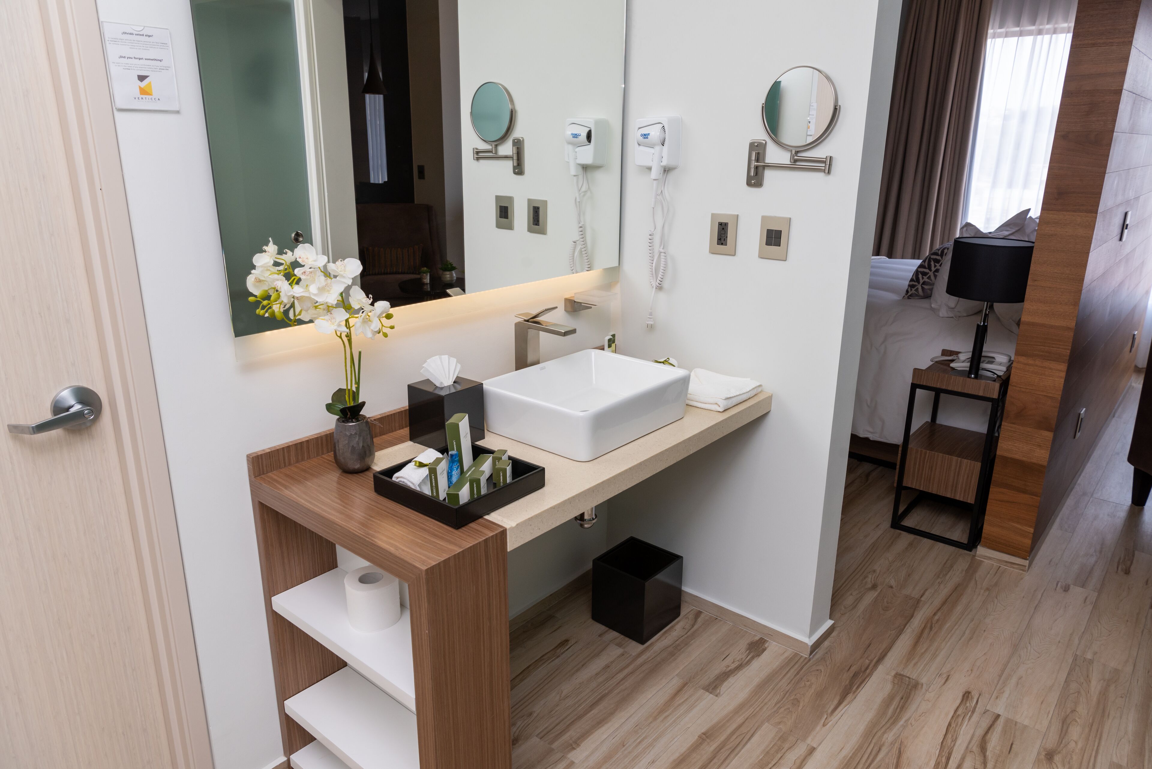 Un baño moderno con un lavamanos blanco, espejo y una mesita de noche con lámpara.