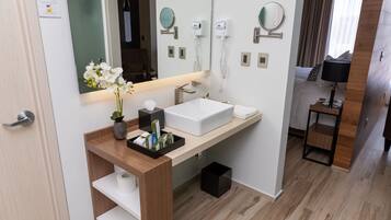 Un baño moderno con un lavamanos blanco, espejo y una mesita de noche con lámpara.