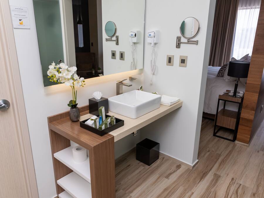 Un baño moderno con un lavamanos blanco, espejo y una mesita de noche con lámpara.