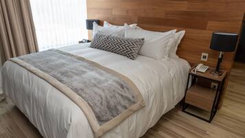 A neatly made bed with a grey throw blanket and a wooden headboard. There is a bedside table with a lamp and a phone.