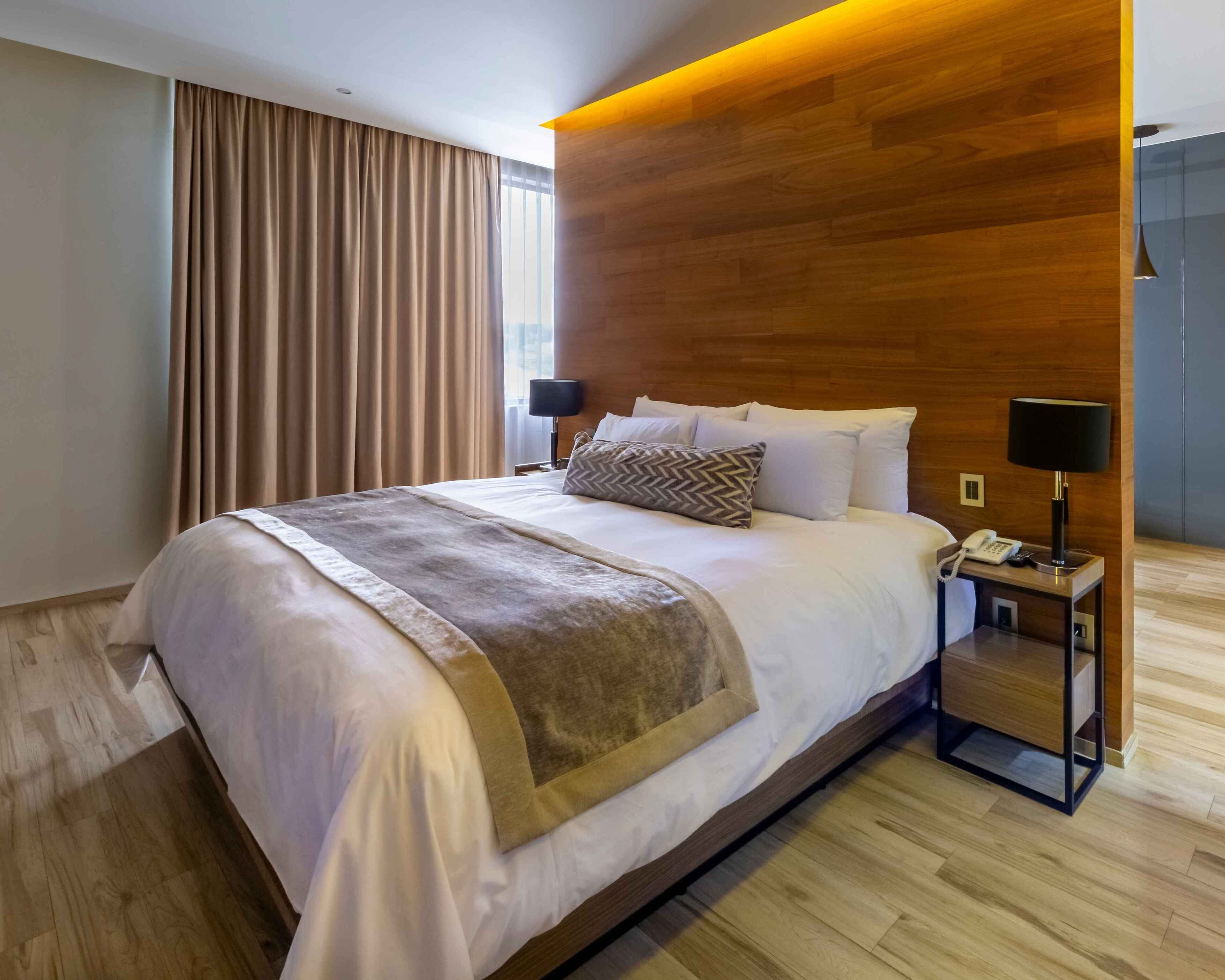 Habitació d'hotel moderna amb un llit gran, un panell de fusta al capçal i una tauleta de nit amb una làmpada.
