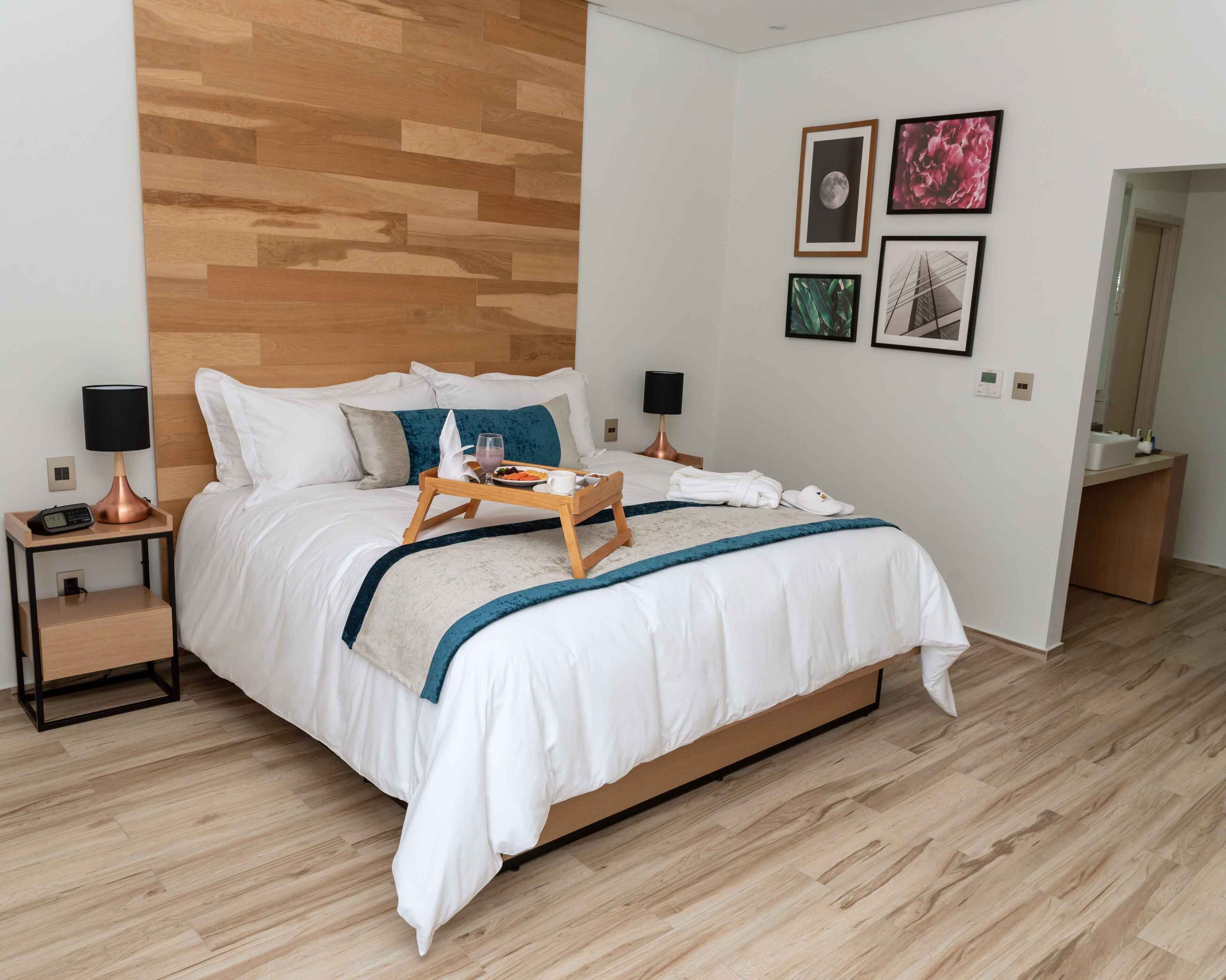 A modern hotel room with a wooden headboard, a neatly made bed with white linens, a bedside table with a lamp, and a tray with breakfast items.
