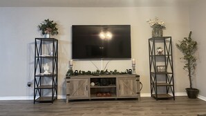 Ein Wohnzimmer mit einem an der Wand montierten Flachbildfernseher, Holzboden und zwei schwarzen Regale mit Pflanzen.