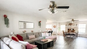 Ein großzügiges Wohnzimmer mit einer großen Eckcouch, einem Couchtisch und einem Essbereich mit Tisch und Stühlen. Im Hintergrund befindet sich ein Deckenventilator und ein Kamin.