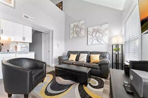 A modern living room with a black leather sofa, a matching armchair, and a coffee table. There are abstract paintings on the wall, a floor lamp, and a window with blinds. - Come to the lake and fish and play! Stay in our Bright Branson 2/2 Condo! (Branson)