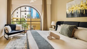 Een hotelkamer met een groot rondboogvenster, een bed met een grijze hoofdeinde, een zwarte fauteuil en een moderne kroonluchter.