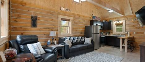 A cozy cabin interior with a wooden interior, a black leather sofa, a dining table, and a kitchen area.