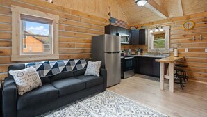 Ein Holzblockhaus-Interieur mit einer Küchenzeile, einem Sofa mit gemusterten Kissen, einem Holzesstisch und einem Fenster mit Blick auf ein Haus.