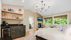 A modern bedroom with a large bed, a TV mounted on the wall, and a view of greenery through the windows. - Avery House in Newberg: 6 bedrooms, forest views, sleeps 12, ideal for family getaways (Newberg)