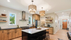 A modern kitchen with wooden cabinets, a large island with stools, pendant lighting, and a view of the outdoors. - Avery House in Newberg: 6 bedrooms, forest views, sleeps 12, ideal for family getaways (Newberg)