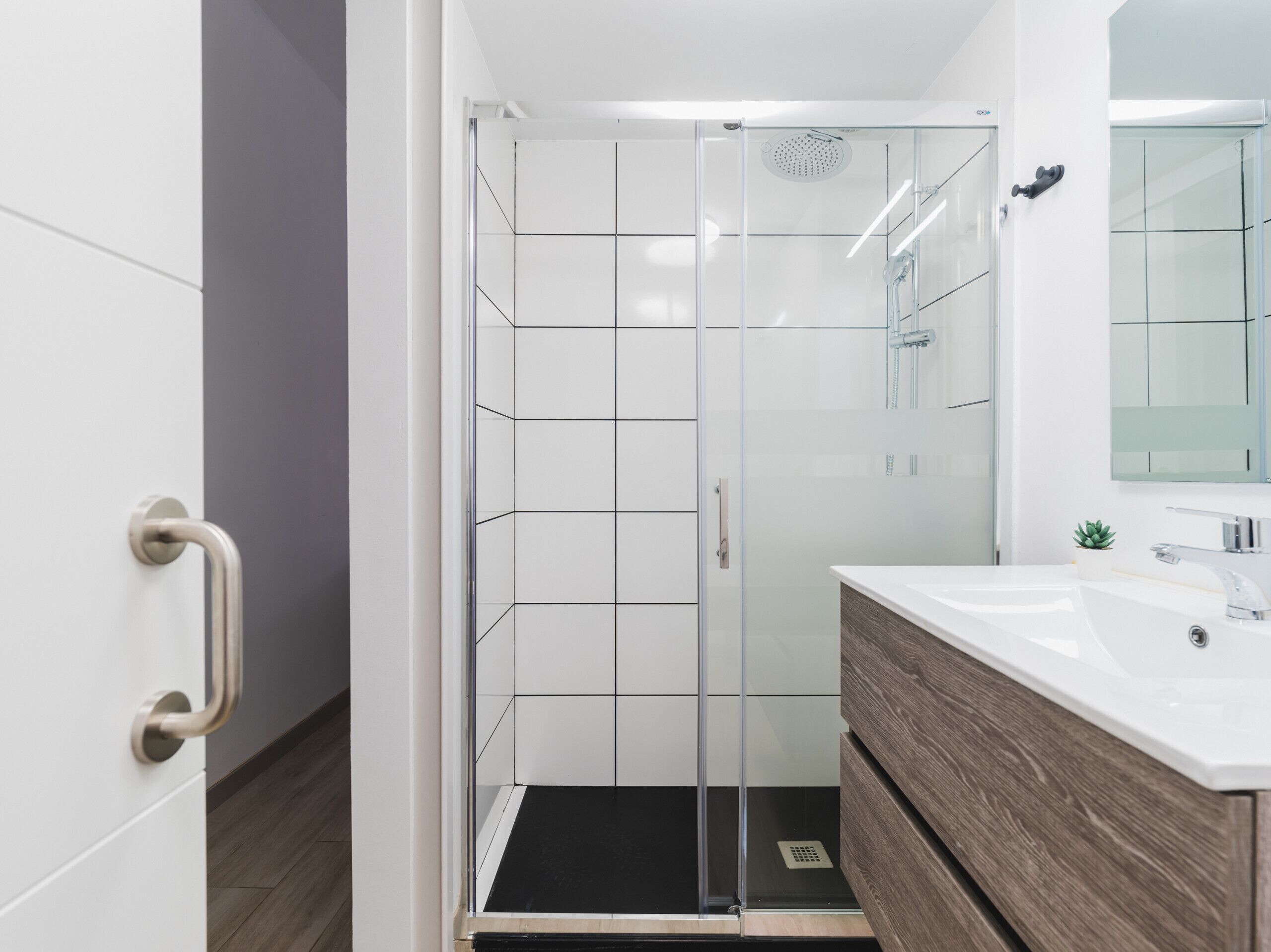 Un baño moderno con cabina de ducha de vidrio, azulejos blancos, un mueble de tocador de madera y un espejo sobre el lavabo.