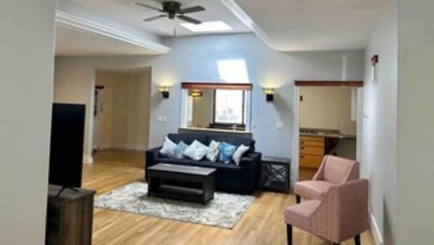A spacious living room with a ceiling fan, a dark-colored sofa with multiple cushions, a small coffee table, and a pink chair.