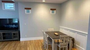 A dining area with a table and chairs, a television in the background, and wooden flooring.