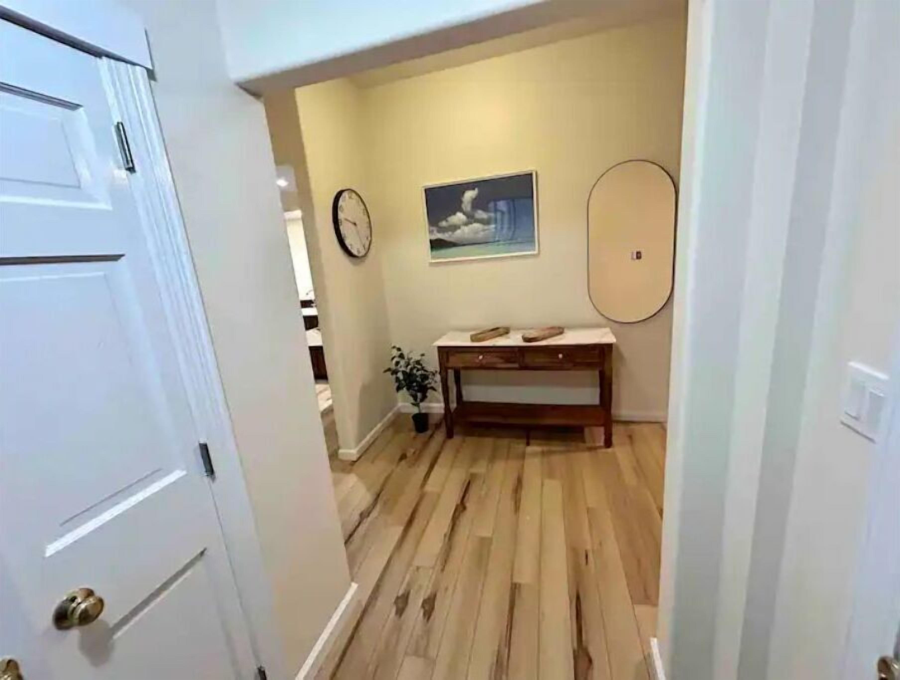 A hallway with wooden flooring, a white door, a clock, a painting, and a small table with a plant.