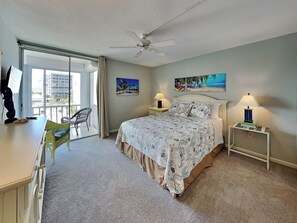 A bedroom with a bed, two bedside tables with lamps, a ceiling fan, a balcony with a chair, and two wall art pieces. - Island Reef 304 by Distinctive Beach Rentals (Fort Myers Beach)