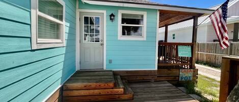Una casa turquesa con terraza de madera, puerta principal y un pequeño porche.