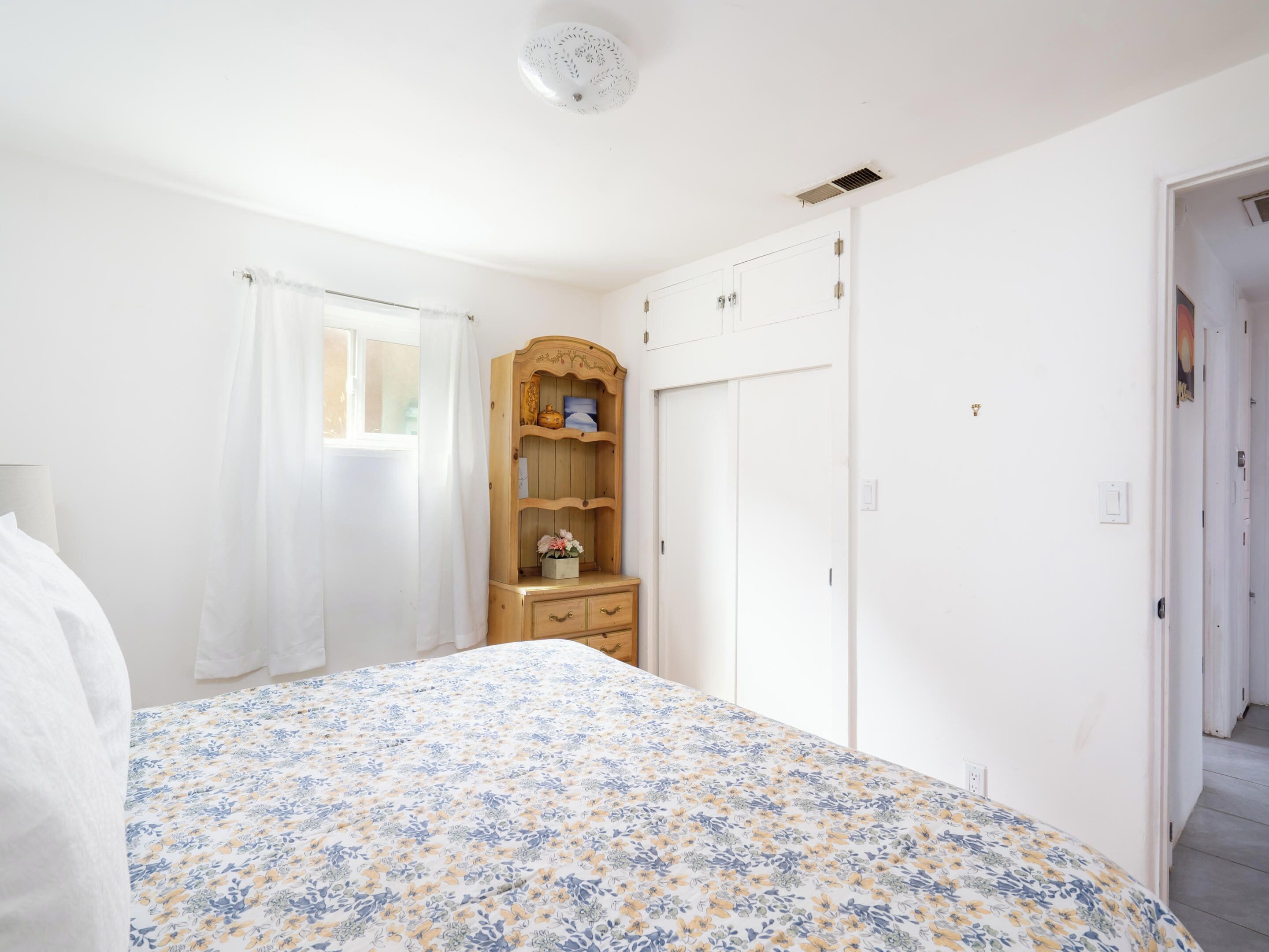 A bedroom with a floral bedspread, a wooden dresser with a mirror, a wardrobe, and a window with white curtains.