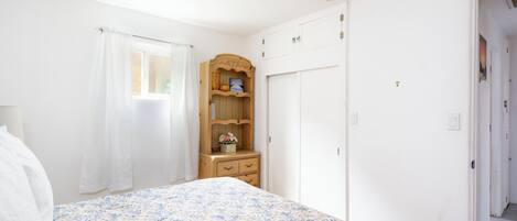 A bedroom with a floral bedspread, a wooden dresser with a mirror, a wardrobe, and a window with white curtains.