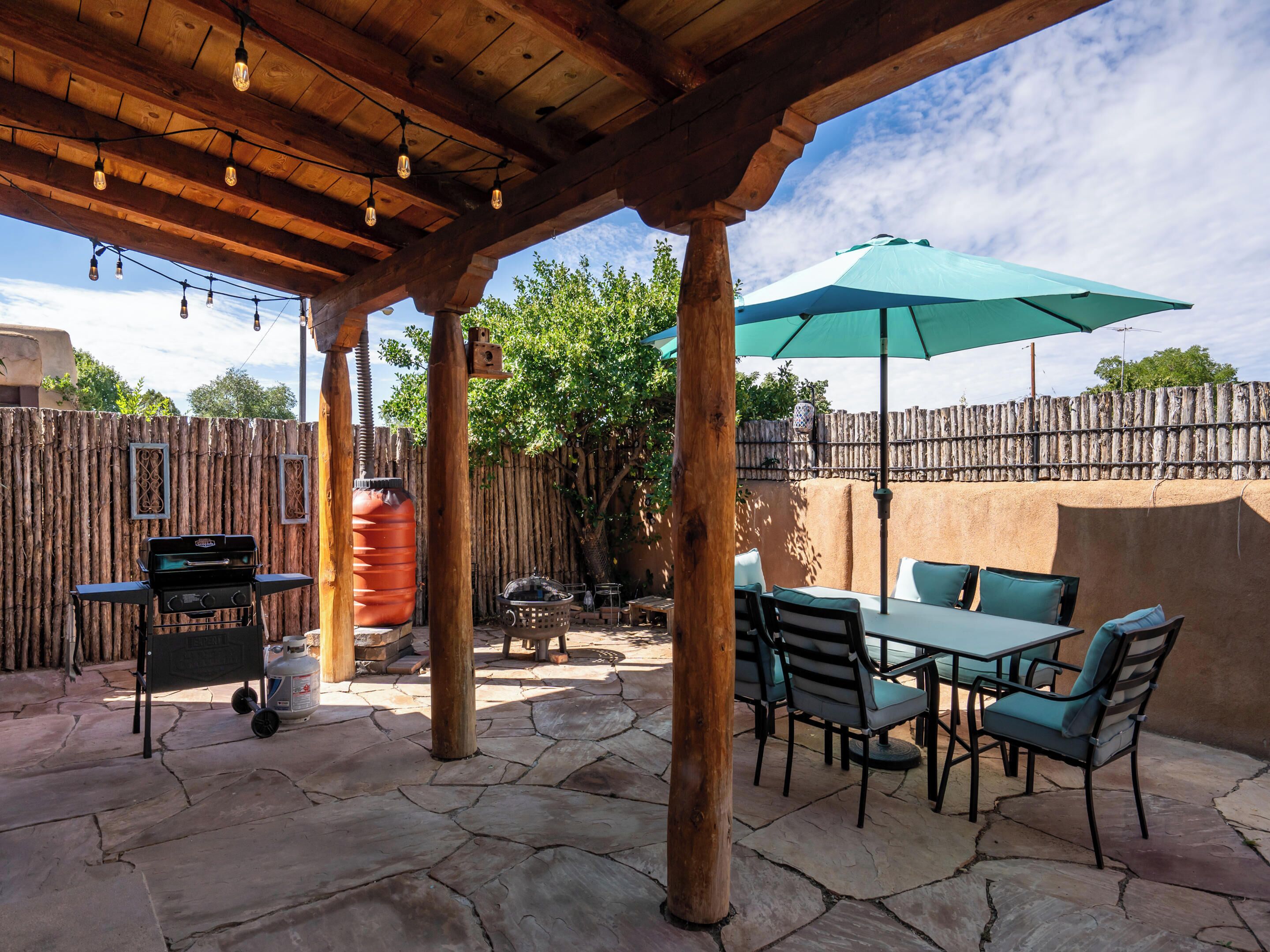 An outdoor patio with stone flooring, wooden pergola, string lights, a table with chairs, and a green umbrella.