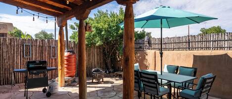 An outdoor patio with stone flooring, wooden pergola, string lights, a table with chairs, and a green umbrella.