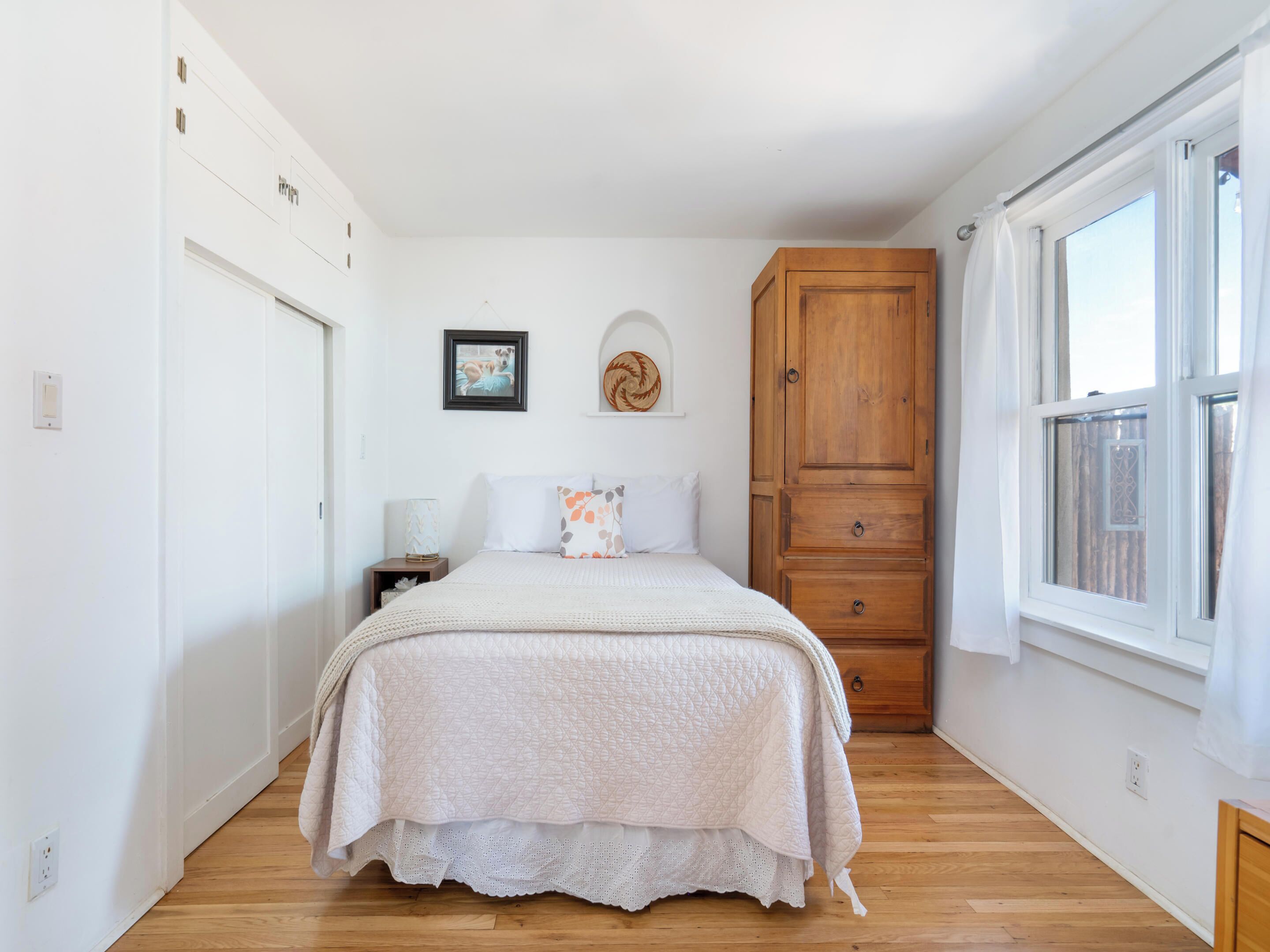 A bedroom with a bed, a wooden wardrobe, a nightstand, a wall-mounted picture, and a window with curtains.