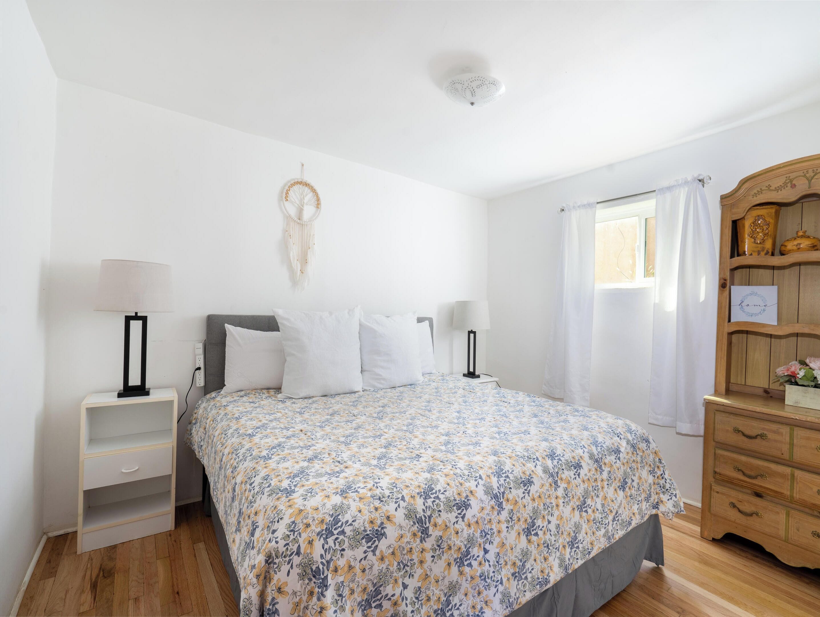 Un dormitorio con colcha estampada, dos mesitas de noche con lámparas, un aparador de madera, un atrapasueños colgado en la pared y una ventana con cortinas blancas.