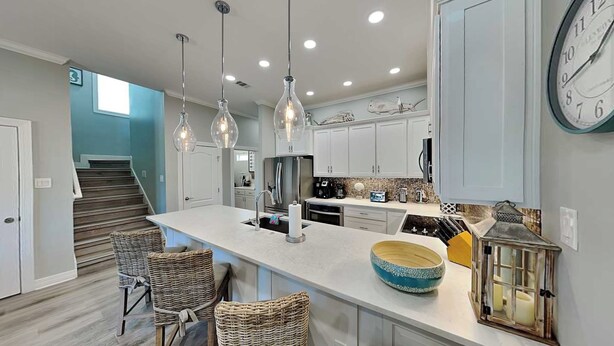 A modern kitchen with a central island, pendant lights, wicker chairs, and a clock on the wall. - Mermaids Lair by Silver Sands Vacation Rentals (Port Aransas)