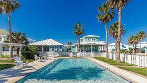 A swimming pool with lounge chairs and palm trees, surrounded by houses with white fences. - Mermaids Lair by Silver Sands Vacation Rentals (Port Aransas)