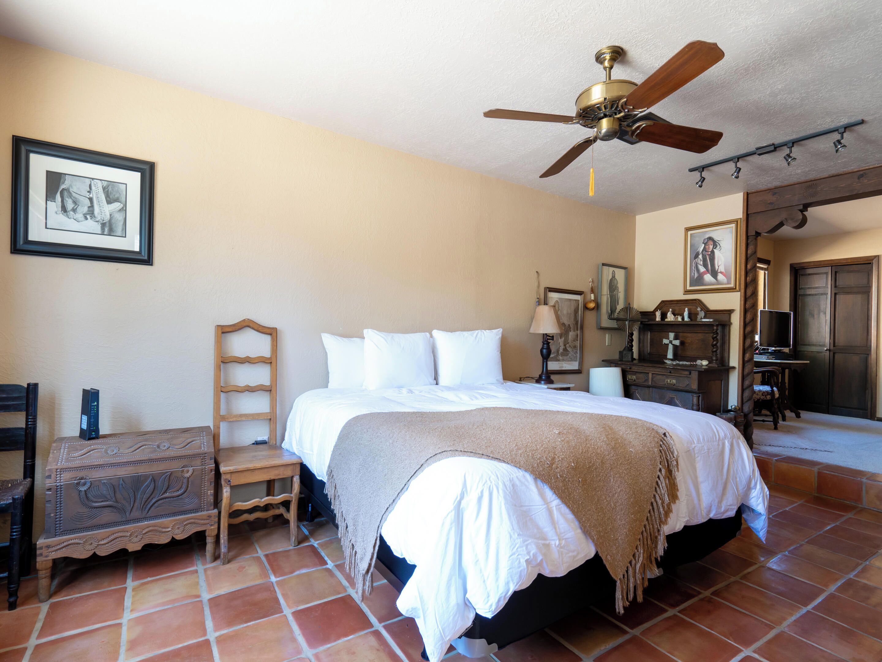 A bedroom with a bed, a wooden nightstand, a chair, a desk, a ceiling fan, and a door.