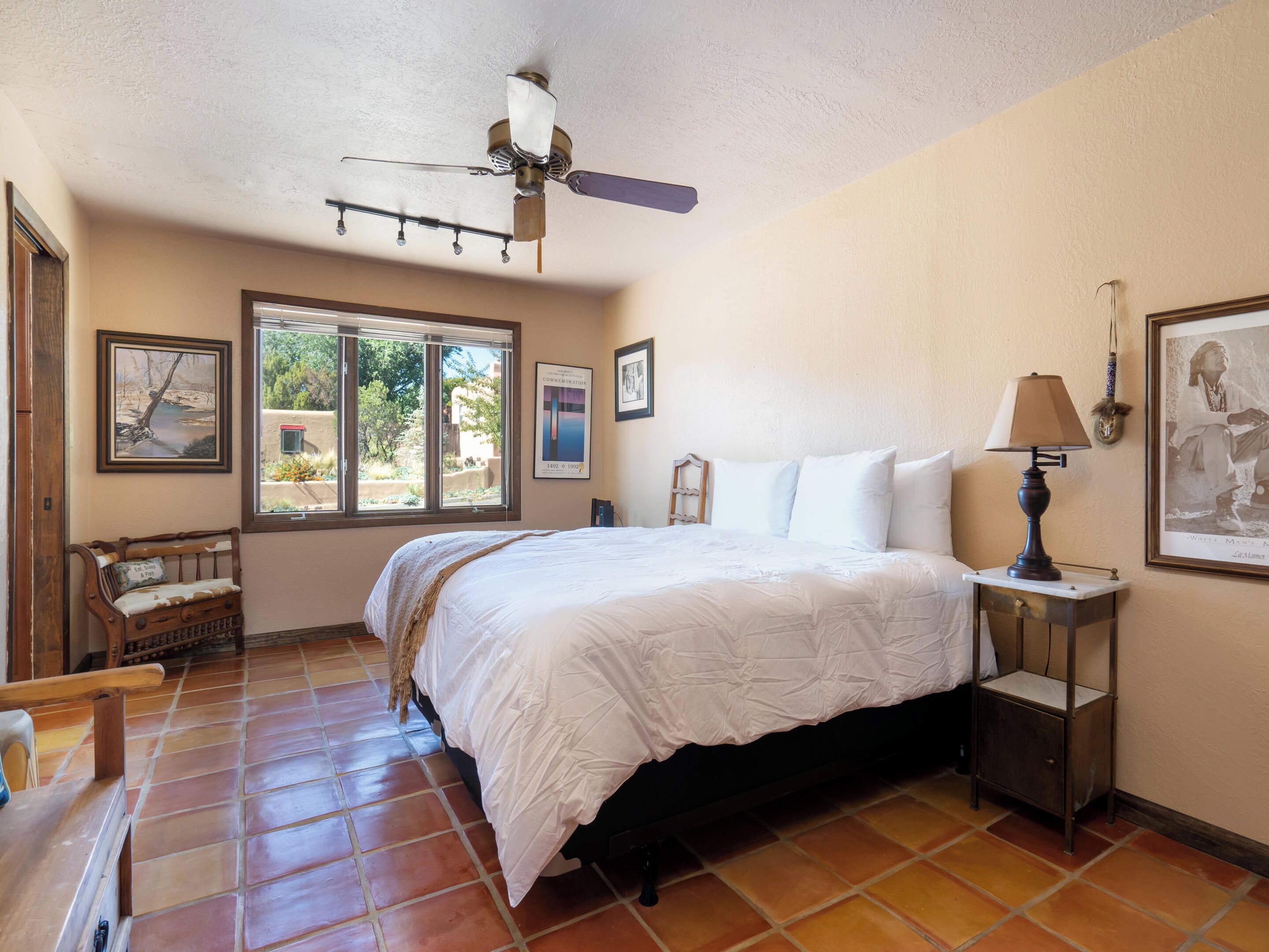 A bedroom with a bed, a nightstand, a lamp, a ceiling fan, a window, and a door.