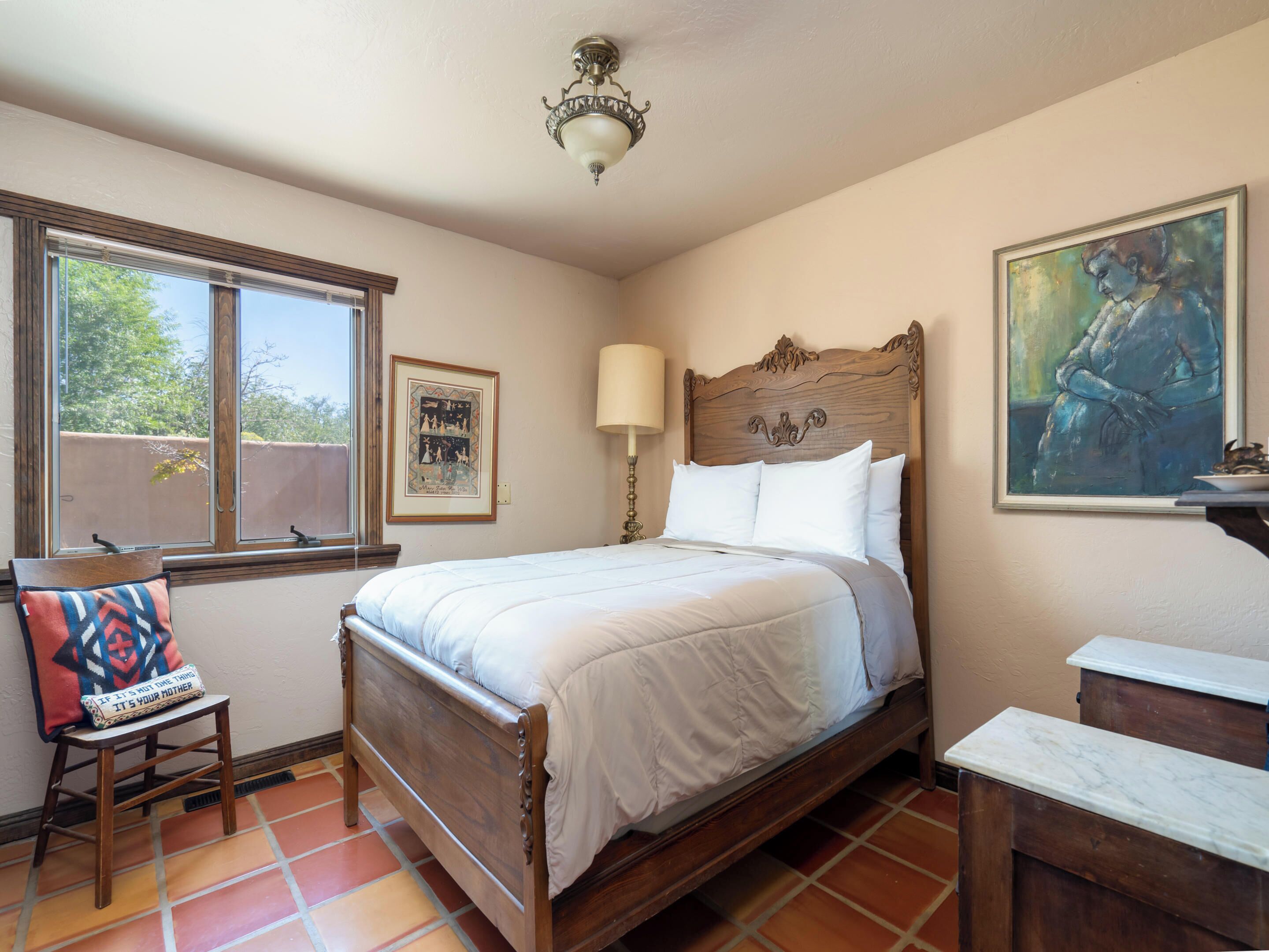 Un dormitorio con cama de madera, una ventana con persianas, una mesita de noche, una silla y un cuadro en la pared.