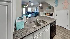 A modern kitchen with granite countertops, a double sink, and a built-in dishwasher. There is a teal-colored toaster and a white cabinet. The room has a dining area with blue chairs and a table. - Trestles by Silver Sands Vacation Rentals (Corpus Christi)