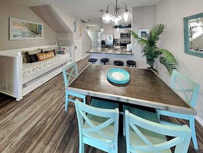 A dining area with a wooden table, four chairs, a blue tray, and a kitchen in the background. - Trestles by Silver Sands Vacation Rentals (Corpus Christi)
