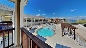 A pool area with a view of the ocean, a wooden deck, and a building with balconies. - Trestles by Silver Sands Vacation Rentals (Corpus Christi)