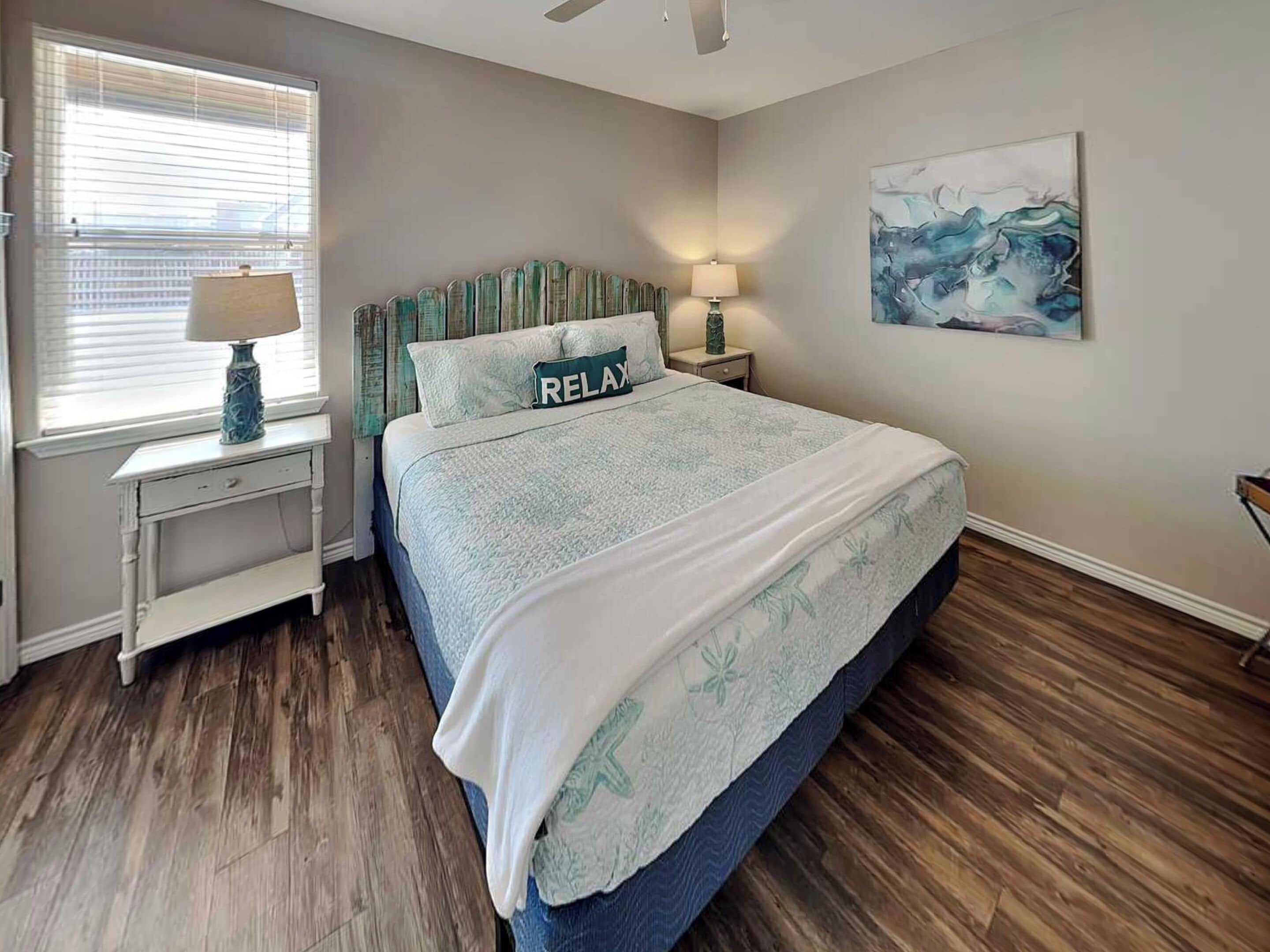 A bedroom with a bed, two bedside tables, a window with blinds, a ceiling fan, and a wall-mounted abstract painting.