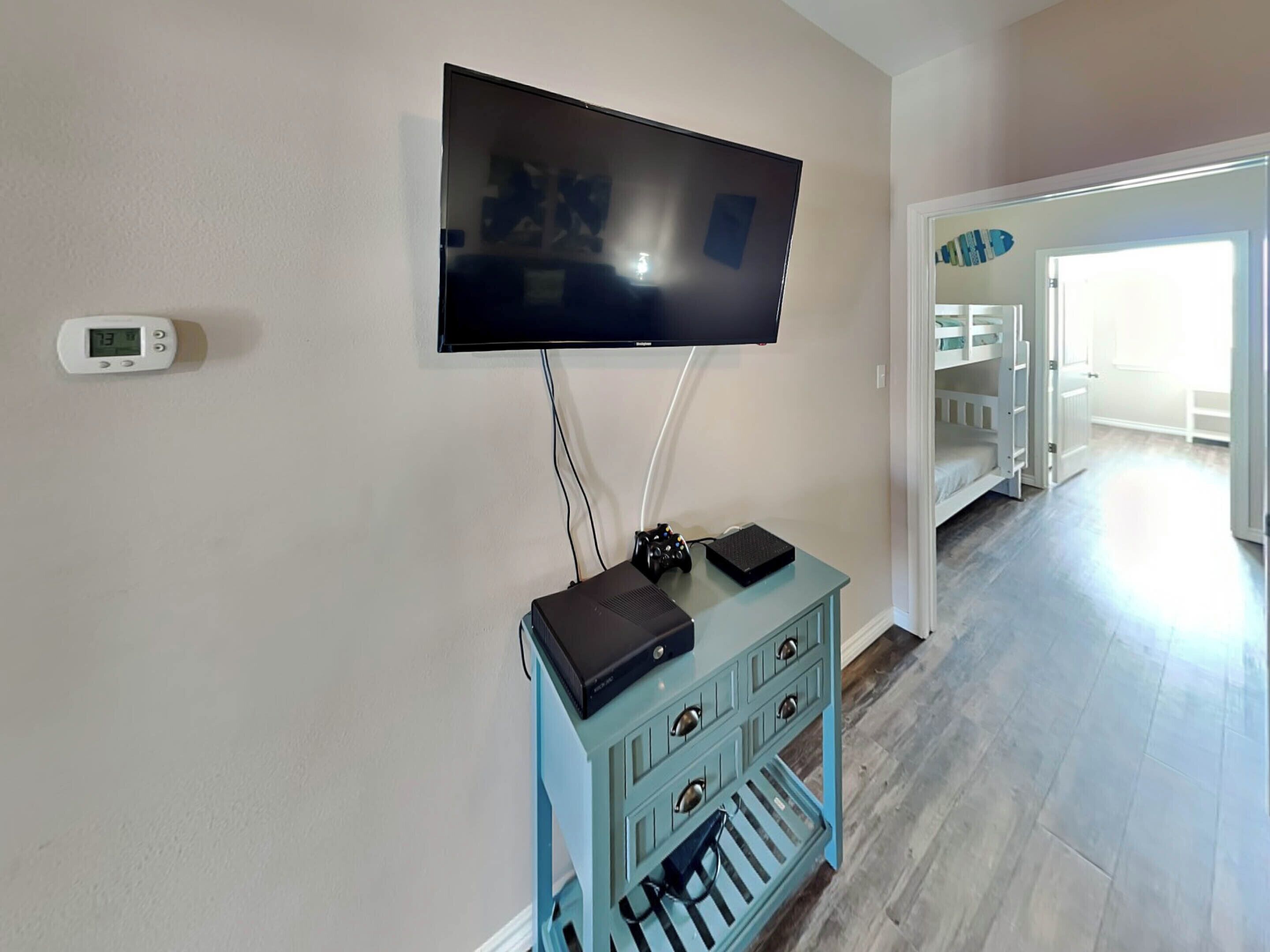 A room with a mounted TV, a digital thermostat, and a blue entertainment unit with a game controller and a black device.