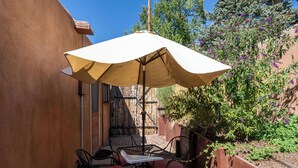 Une terrasse avec une table et des chaises, protégée par un grand parasol, entourée d’une clôture en bois et d’un jardin où poussent des fleurs violettes.