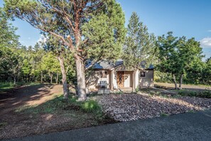 A single-story house with a sloped roof, surrounded by trees and a gravel driveway.